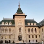 Grand beige city-building with a central tower, green roofs, and arched windows against a clear blue sky.