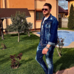 Man in a denim jacket and jeans stands on a grassy lawn beside a blue swimming pool in a suburban yard with an orange house in the background, wearing sunglasses.