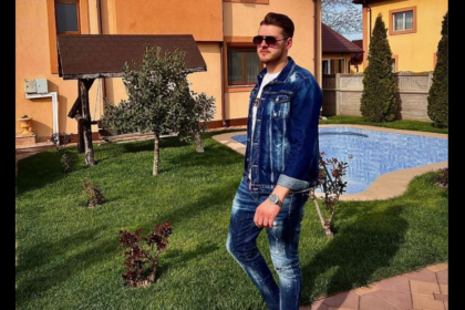 Man in a denim jacket and jeans stands on a grassy lawn beside a blue swimming pool in a suburban yard with an orange house in the background, wearing sunglasses.