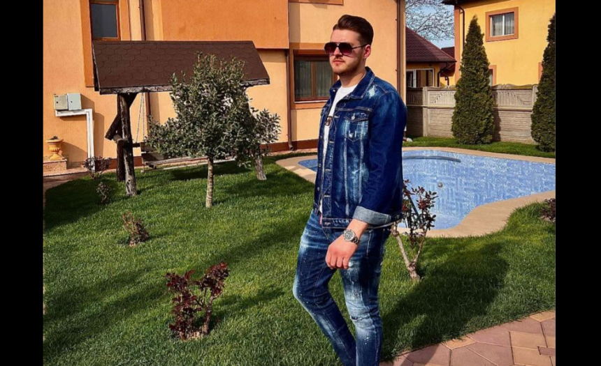 Man in a denim jacket and jeans stands on a grassy lawn beside a blue swimming pool in a suburban yard with an orange house in the background, wearing sunglasses.