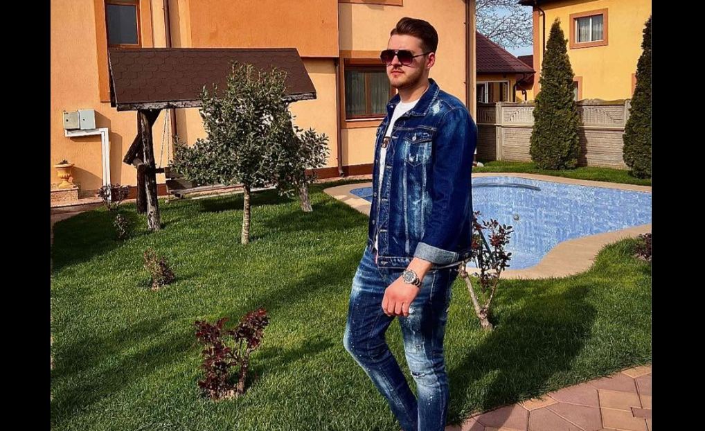 Man in a denim jacket and jeans stands on a grassy lawn beside a blue swimming pool in a suburban yard with an orange house in the background, wearing sunglasses.