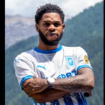 Athlete posing outdoors with mountains in the background, arms crossed, wearing a white and blue soccer jersey with a crest on the chest.