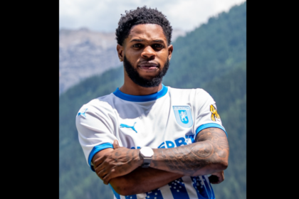 Athlete posing outdoors with mountains in the background, arms crossed, wearing a white and blue soccer jersey with a crest on the chest.