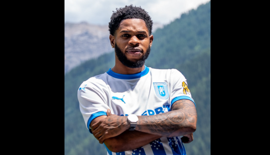 Athlete posing outdoors with mountains in the background, arms crossed, wearing a white and blue soccer jersey with a crest on the chest.