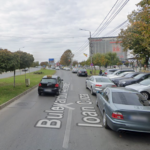 Two-lane urban street with cars parked along the right curb, a grassy median with trees on the left, and a storefront building with a Kik sign in the distance.