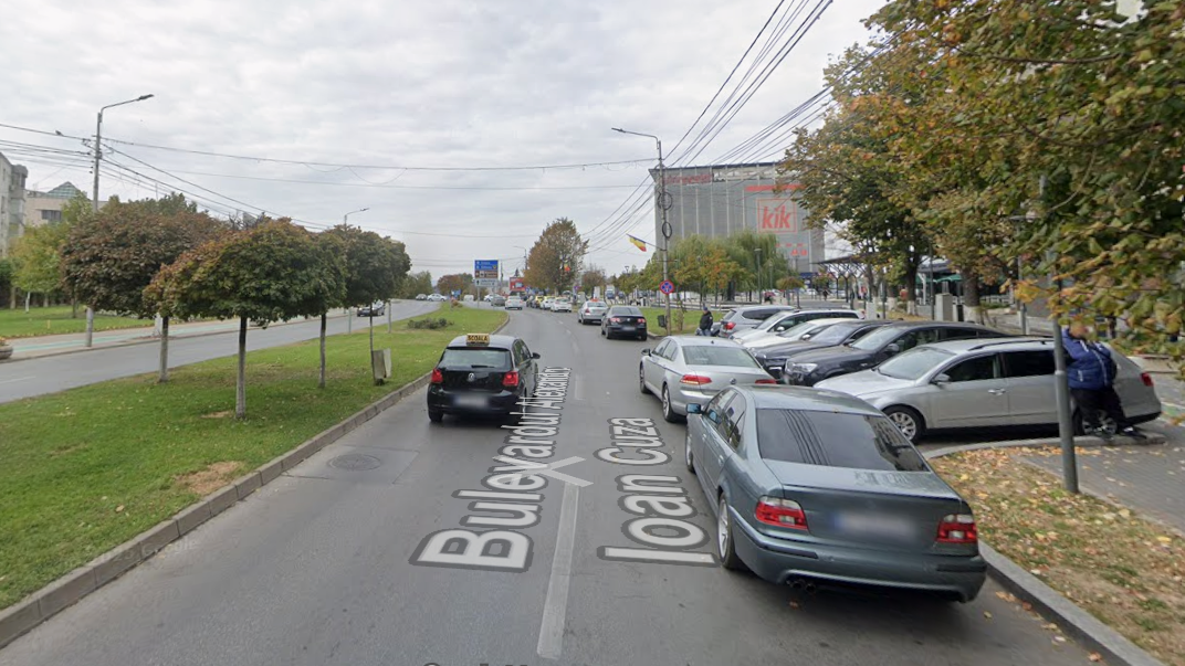 Two-lane urban street with cars parked along the right curb, a grassy median with trees on the left, and a storefront building with a Kik sign in the distance.