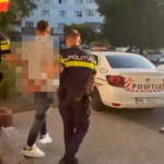 Three police officers in dark uniforms with yellow stripes detain a pixelated individual outside a building, beside a white police car labeled POLITIA. A residential area is in the background.