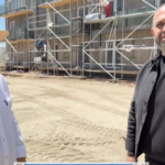 Two men stand at a construction site with scaffolding in the background; one wears a white shirt and straw hat, the other a black shirt/jacket on a sunny day?