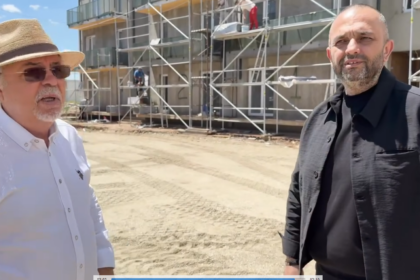 Two men stand at a construction site with scaffolding in the background; one wears a white shirt and straw hat, the other a black shirt/jacket on a sunny day?