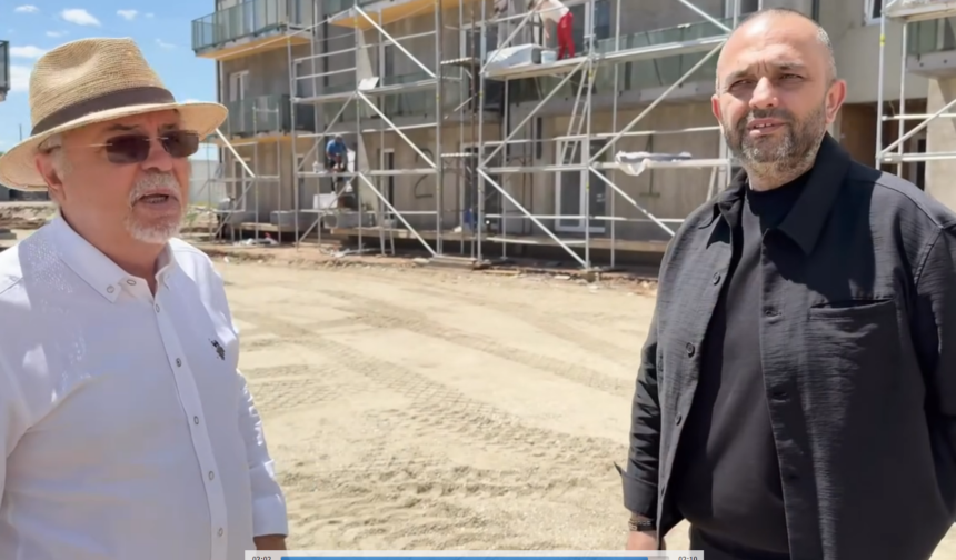 Two men stand at a construction site with scaffolding in the background; one wears a white shirt and straw hat, the other a black shirt/jacket on a sunny day?