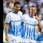 Two soccer players in white-and-blue striped jerseys stand together on the field, smiling at the camera; one shows a thumbs-up gesture.