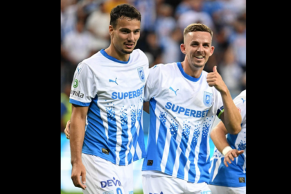 Two soccer players in white-and-blue striped jerseys stand together on the field, smiling at the camera; one shows a thumbs-up gesture.