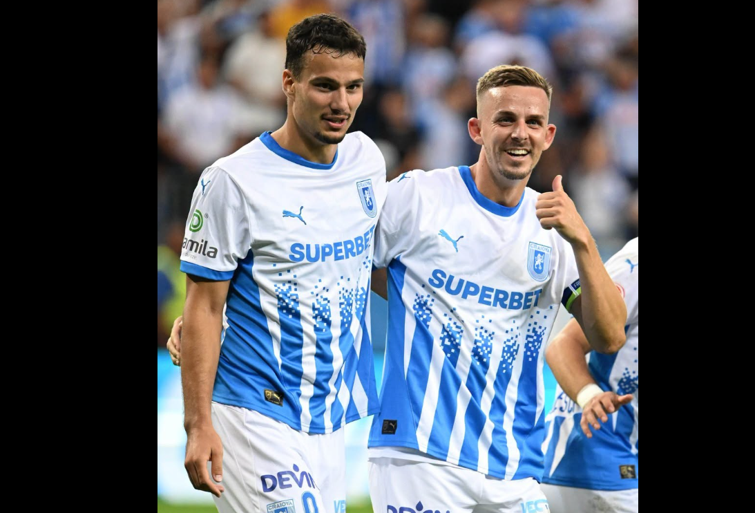 Two soccer players in white-and-blue striped jerseys stand together on the field, smiling at the camera; one shows a thumbs-up gesture.