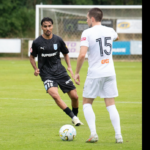 Two soccer players compete for the ball on a green field, one in a black kit and the other in white with the number 15 on his back, near a goal.