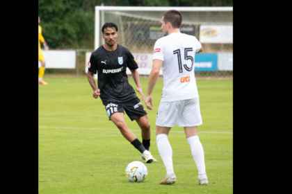 Two soccer players compete for the ball on a green field, one in a black kit and the other in white with the number 15 on his back, near a goal.