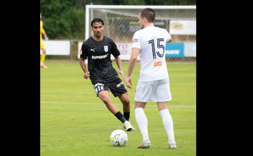 Two soccer players compete for the ball on a green field, one in a black kit and the other in white with the number 15 on his back, near a goal.