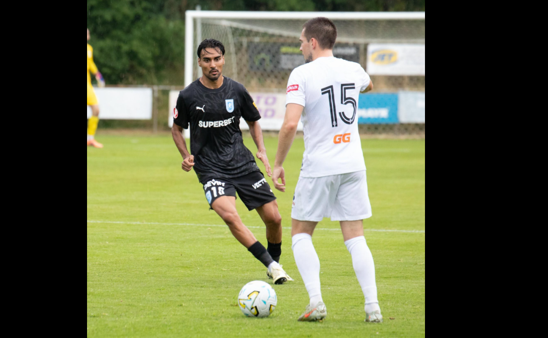 Two soccer players compete for the ball on a green field, one in a black kit and the other in white with the number 15 on his back, near a goal.