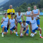 Soccer team posing on a green field; goalkeeper in yellow stands behind, players in white jerseys and blue shorts.