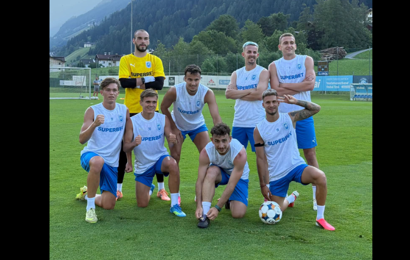 Soccer team posing on a green field; goalkeeper in yellow stands behind, players in white jerseys and blue shorts.