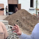 Two men converse at a construction site, with a large dirt heap and a building in the background. One wears a baseball cap and tan polo; the other wears a straw hat, sunglasses, and a white shirt.