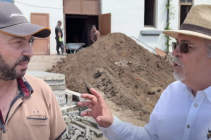 Two men converse at a construction site, with a large dirt heap and a building in the background. One wears a baseball cap and tan polo; the other wears a straw hat, sunglasses, and a white shirt.