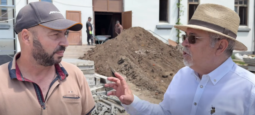 Two men converse at a construction site, with a large dirt heap and a building in the background. One wears a baseball cap and tan polo; the other wears a straw hat, sunglasses, and a white shirt.