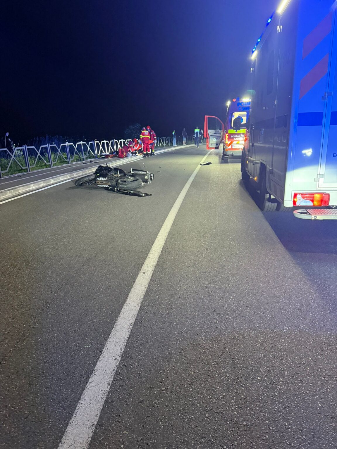 Nighttime road accident scene with a damaged Yamaha motorcycle on the road and emergency responders nearby near a barrier fence on the left side of the road.