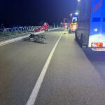 Nighttime road accident scene with a damaged Yamaha motorcycle on the road and emergency responders nearby near a barrier fence on the left side of the road.