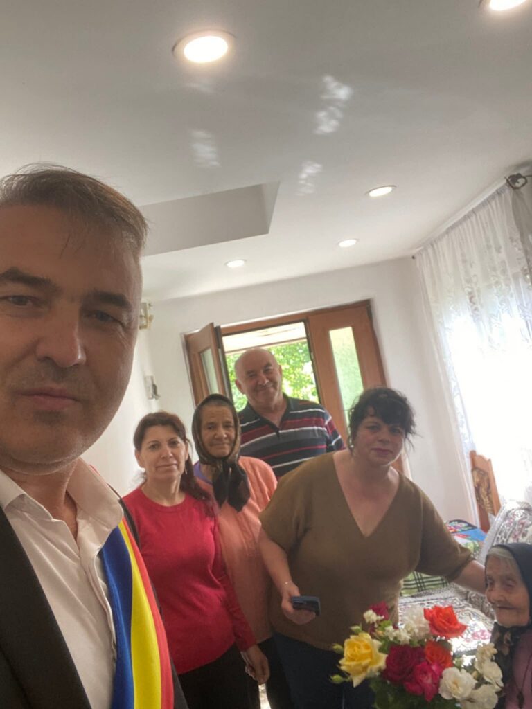 Group of adults posing for a selfie in a bright living room, with a vase of colorful flowers on the table nearby.