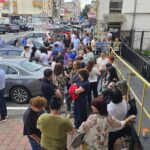 Crowd of people gathered on a sidewalk and ramp outside a building on a busy city street, many talking in groups.