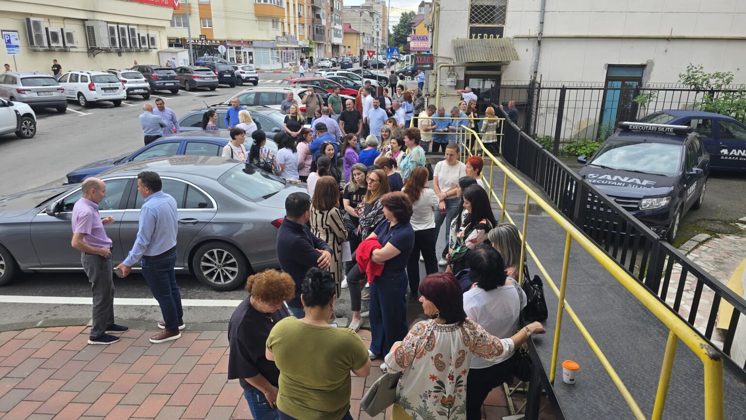 Crowd of people gathered on a sidewalk and ramp outside a building on a busy city street, many talking in groups.