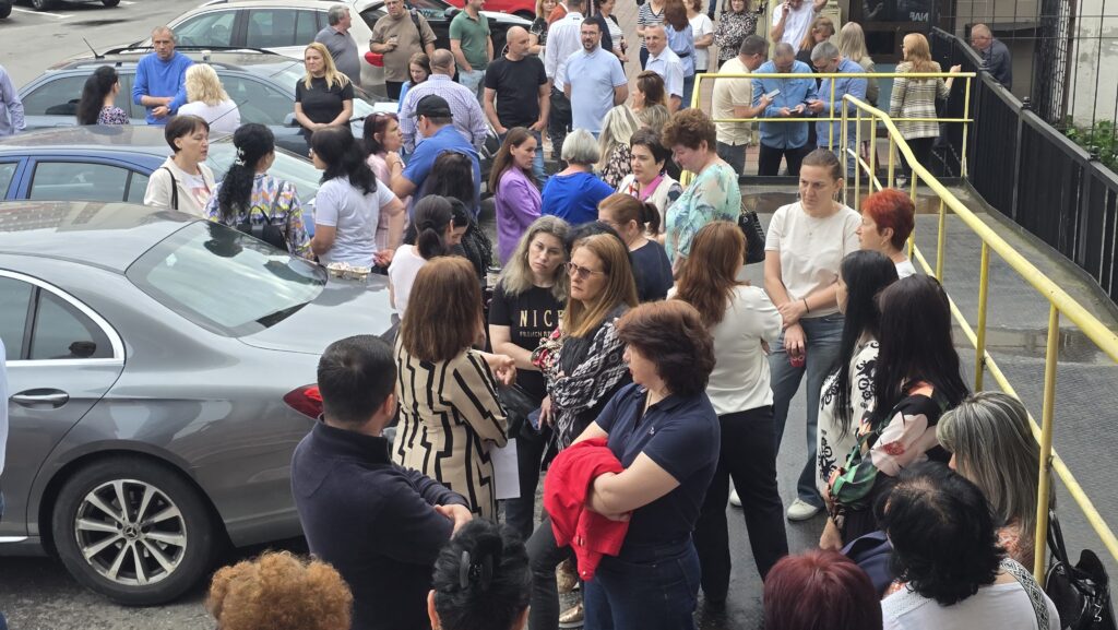 A crowd of adults standing and chatting around a row of parked cars in a parking lot, with a yellow railing and a building in the background.