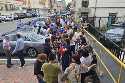 Crowd of people gathered on a sidewalk and ramp outside a building on a busy city street, many talking in groups.