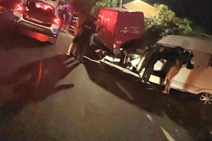 Nighttime car crash on a street with several damaged vehicles and people assisting near the wreckage under streetlights, with a red van involved in the collision.