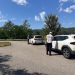 Two police officers check a white SUV in a parking lot, with a second car nearby and a rural, tree-lined background on a sunny day