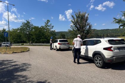 Two police officers check a white SUV in a parking lot, with a second car nearby and a rural, tree-lined background on a sunny day