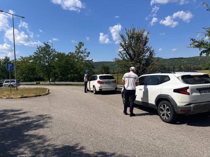 Two police officers check a white SUV in a parking lot, with a second car nearby and a rural, tree-lined background on a sunny day