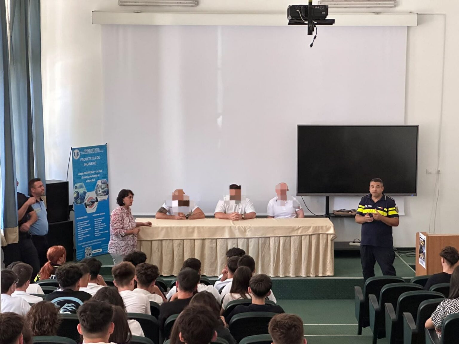 Lecție de viață: deținuți față în față cu elevii la Târgu Jiu - Jurnalul Olteniei – Știri din Craiova și Oltenia Panel of four people seated behind a long table on a stage in a school auditorium, audience of students in front; a uniformed man speaks at right.