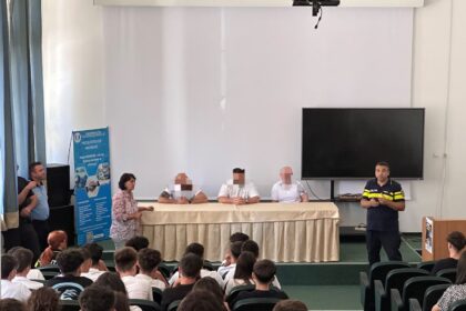 Panel of four people seated behind a long table on a stage in a school auditorium, audience of students in front; a uniformed man speaks at right.