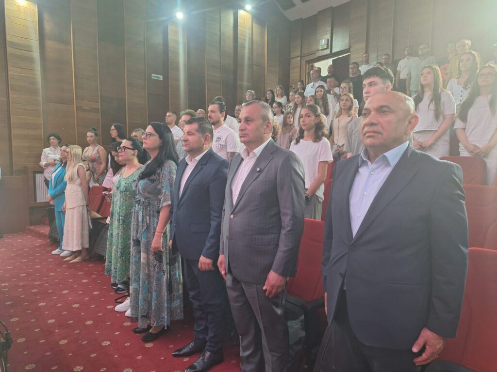 WhatsApp Image 2026-06-09 at 134458 1 - Jurnalul Olteniei – Știri din Craiova și Oltenia Officials in suits stand at attention in a red-carpeted auditorium, with students standing in bleachers behind them and wood-paneled walls.