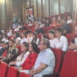 Audience seated in red theater chairs, watching an event; mix of students and adults in a lecture-room setting.