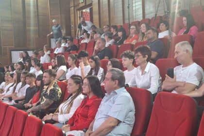 Audience seated in red theater chairs, watching an event; mix of students and adults in a lecture-room setting.