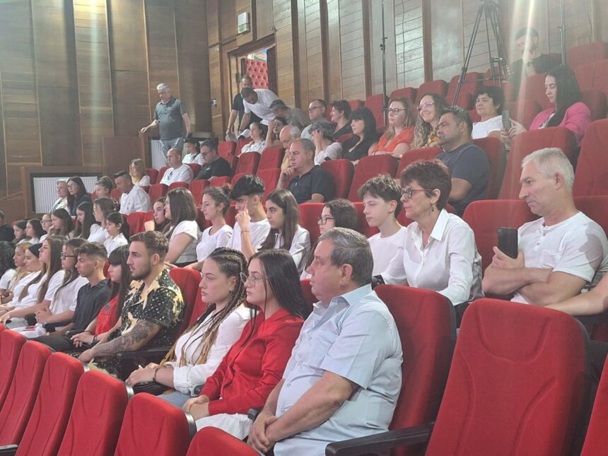 Audience seated in red theater chairs, watching an event; mix of students and adults in a lecture-room setting.