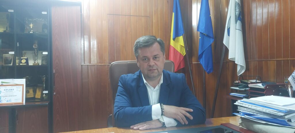 WhatsApp Image 2026-06-11 at 145656 1 - Jurnalul Olteniei – Știri din Craiova și Oltenia Man in a blue suit seated at a desk in a wood-paneled office with flags behind him (Romania, EU, and an additional flag).