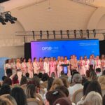 Group of women on a stage at a conference, standing in a row in front of a blue backdrop with OFSD Dolj branding. Audience seated in front.