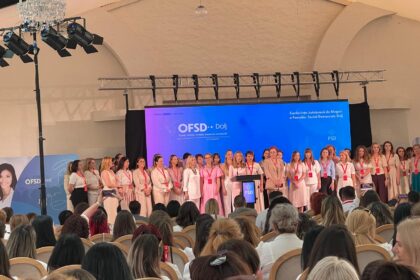Group of women on a stage at a conference, standing in a row in front of a blue backdrop with OFSD Dolj branding. Audience seated in front.