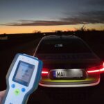 Handheld breathalyzer device held in front of a police car at sunset; the screen shows a 0.0 mg/L reading.