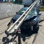 Blue car crashed into a broken concrete utility pole on a street, front severely damaged and shattered windshield, debris scattered nearby.