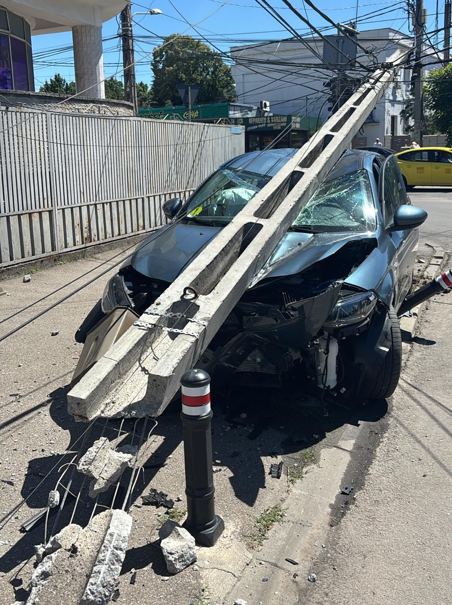WhatsApp Image 2026-06-16 at 153050 - Jurnalul Olteniei – Știri din Craiova și Oltenia Blue car crashed into a broken concrete utility pole on a street, front severely damaged and shattered windshield, debris scattered nearby.