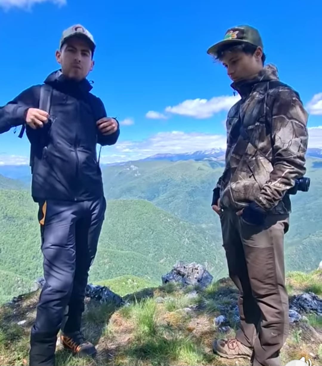 Two hikers stand on a rocky mountain edge, backpacks on, with green valleys and distant snow-capped peaks under a bright blue sky.